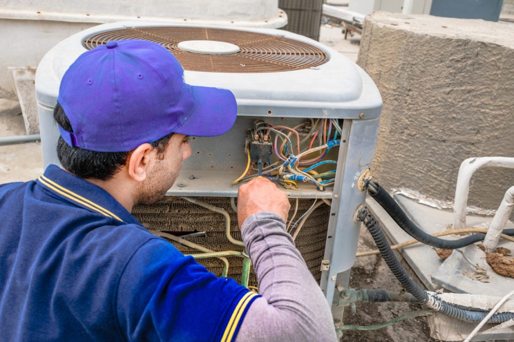 a professional electrician man is giving up thumbs up after the fixing a heavy duty air conditioner on the roof top and wearing blue uniform and cap and the city is in the background