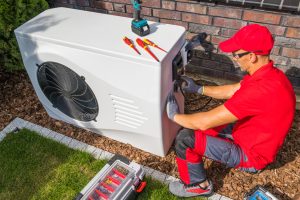 Professional Middle Aged HVAC Technician in Red Uniform Repairing Modern Heat Pump Unit.